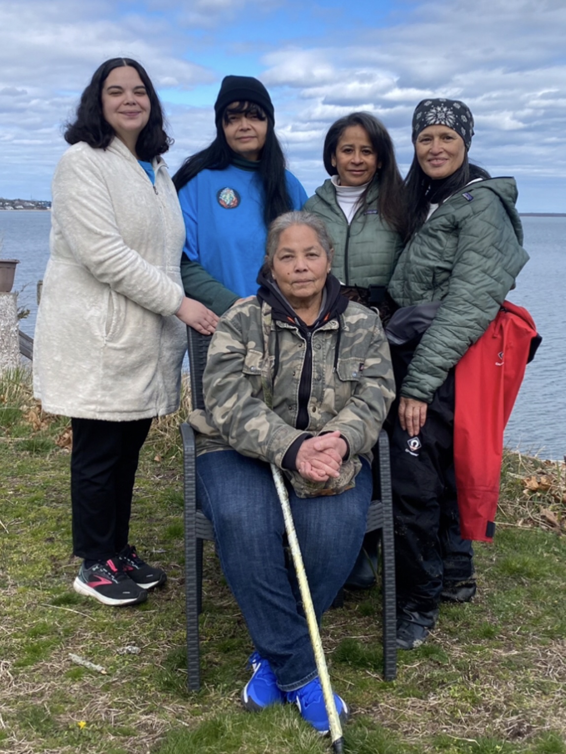 Shinnecock Kelp Farmers Five Shinnecock Farmers posing together outdoors, with a serene lake and cloudy sky in the background. One person is seated in the front, holding a cane, while four others stand behind, smiling.