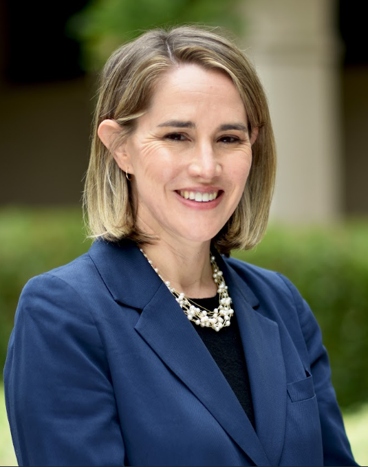 Sarah Reissman Person smiling, wearing a blue blazer and a decorative necklace, standing outdoors.