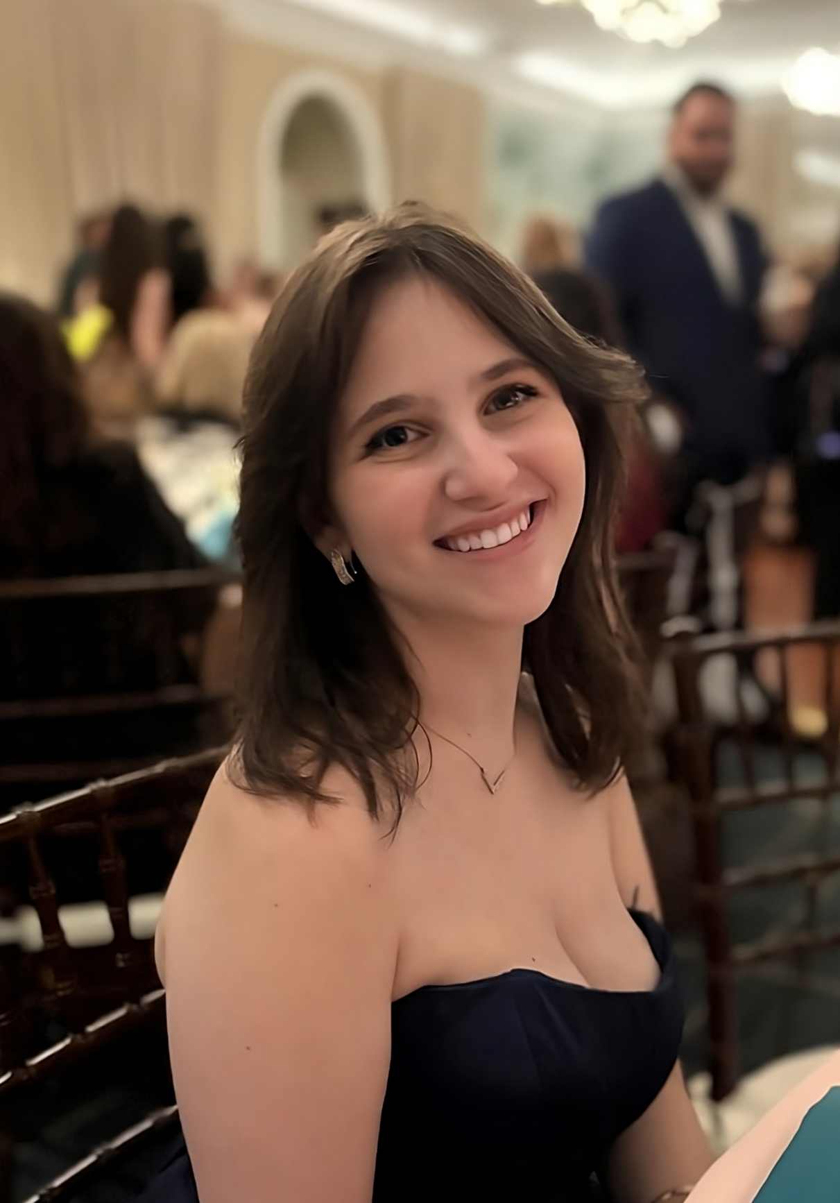Person smiling at a formal event inside an elegant room with other guests in the background.