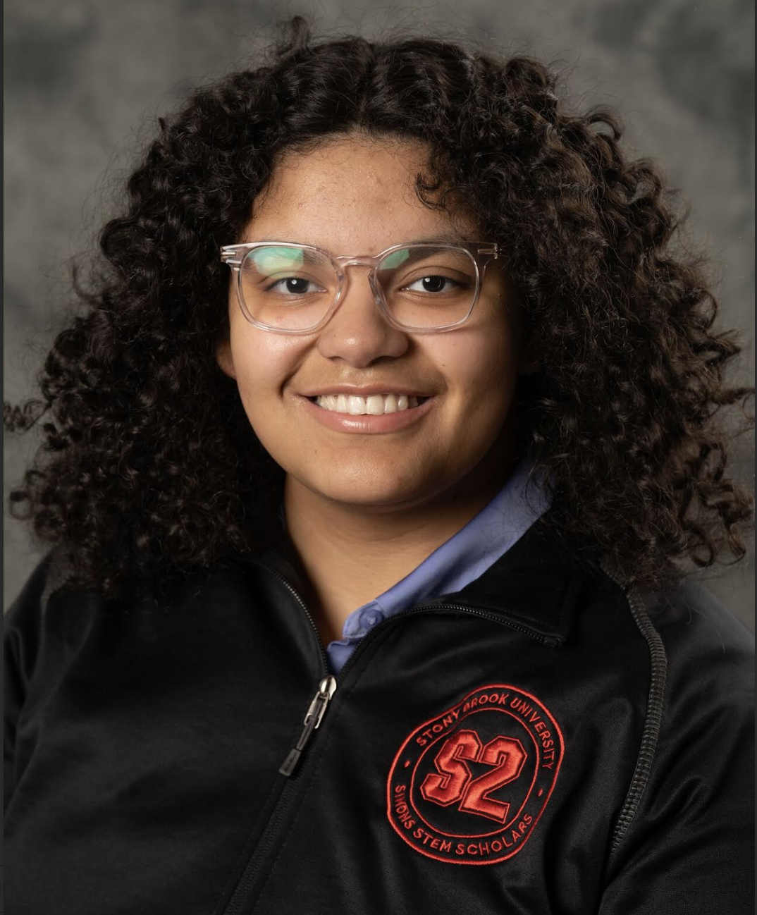 Portrait of a Stony Brook University System Scholar wearing glasses and a dark jacket with the university logo. The individual has curly hair and is smiling at the camera.