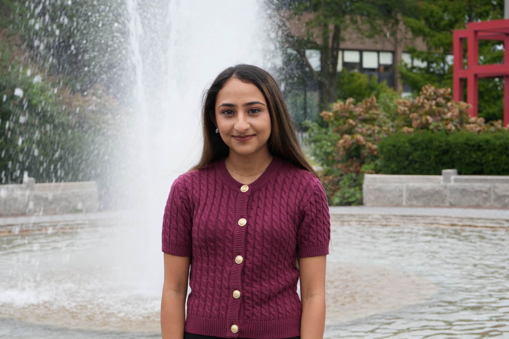A person in a burgundy cardigan stands smiling in front of a fountain.