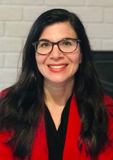 Sheri Levy A woman with long dark hair and glasses smiles warmly, wearing a red jacket. She stands in front of a white brick wall, conveying a friendly and professional tone.