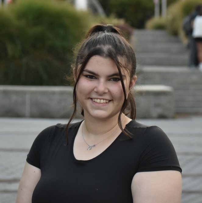 Sophia Goodkin Portrait of a smiling person with pulled back hair, wearing a black top and a necklace, outdoors.