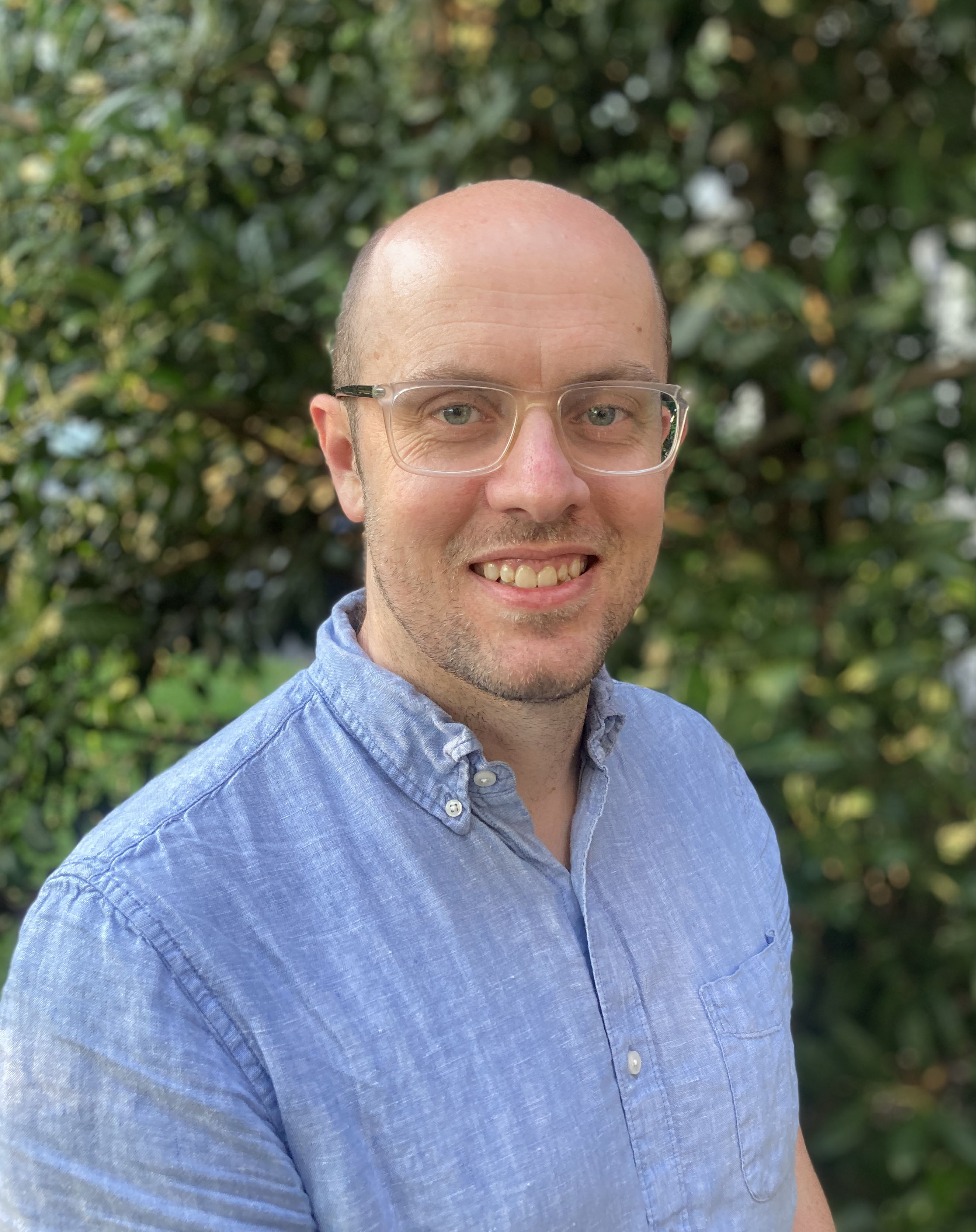 Male wearing clear glasses and blue chambray poses in front of greenery 
