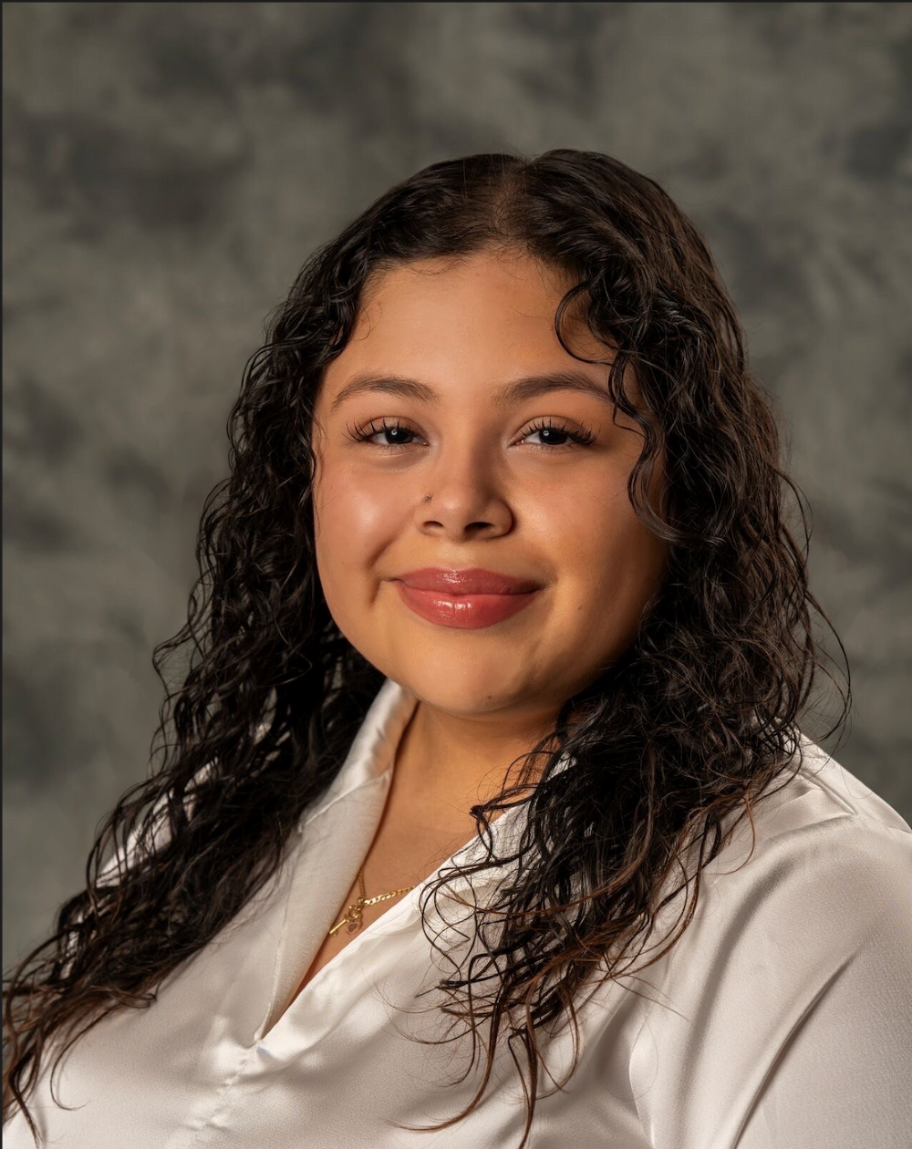 Portrait of a smiling individual with curly hair, wearing a white shirt against a mottled gray background.