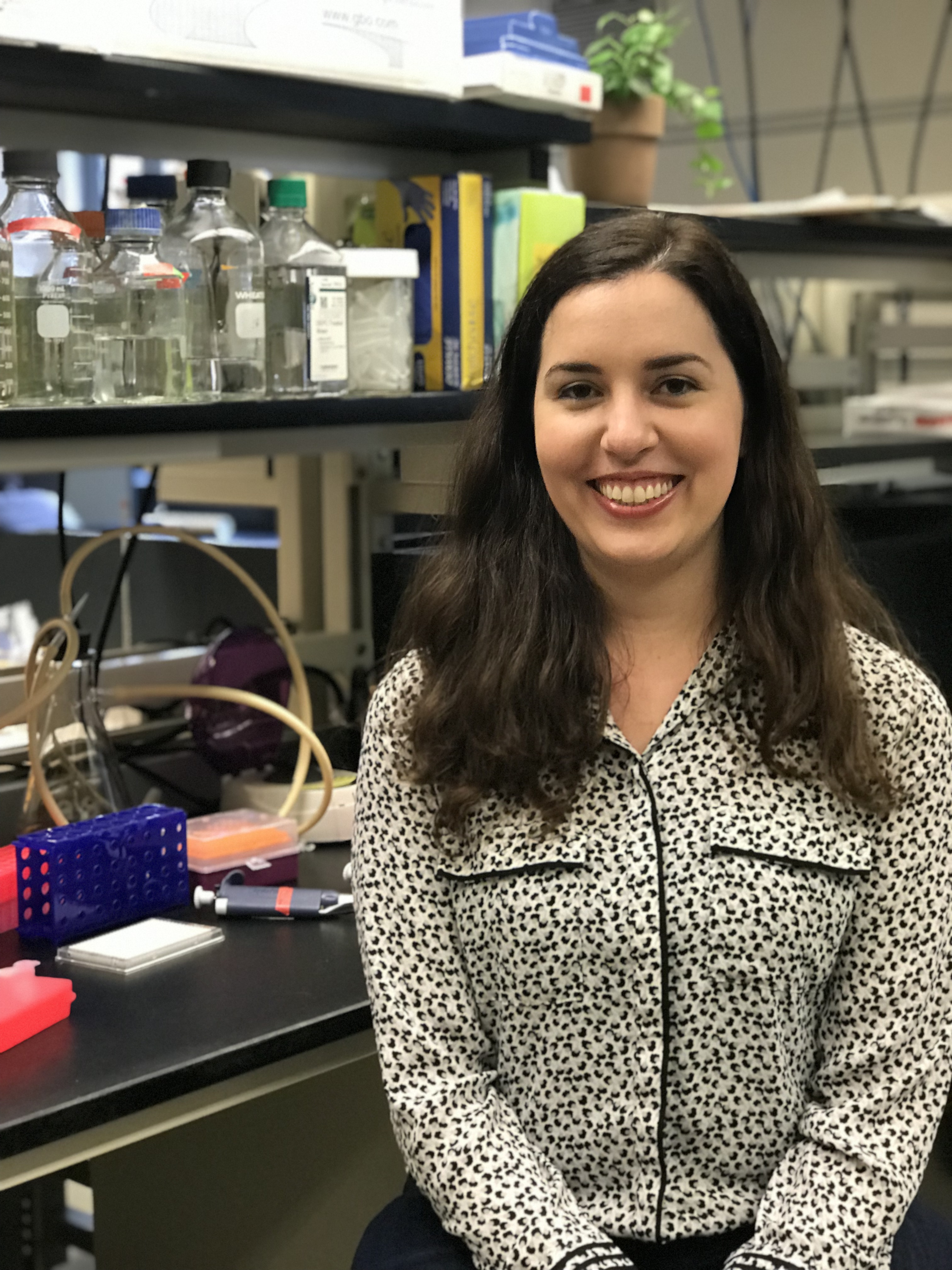 A person smiling in a laboratory with various scientific equipment and supplies in the background.