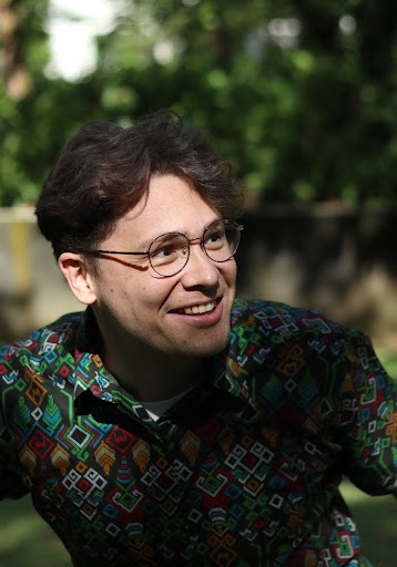 Person in a vibrant geometric-patterned shirt and round glasses smiling outdoors with a blurred green background.