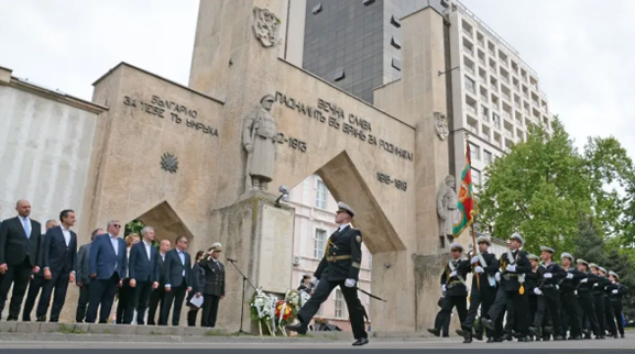 Ceremonial military parade in front of a stone monument, with officials in suits watching. Soldiers in navy uniforms march solemnly, conveying respect.