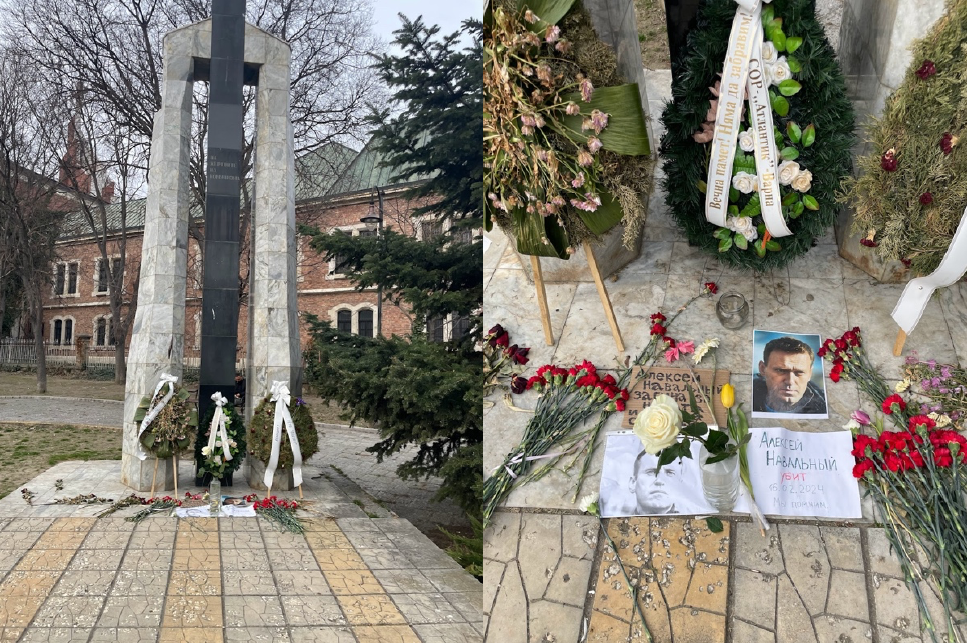 A memorial site with a tall stone monument flanked by white floral wreaths and ribbons. Flowers and photos are placed at the base, conveying respect and remembrance.
