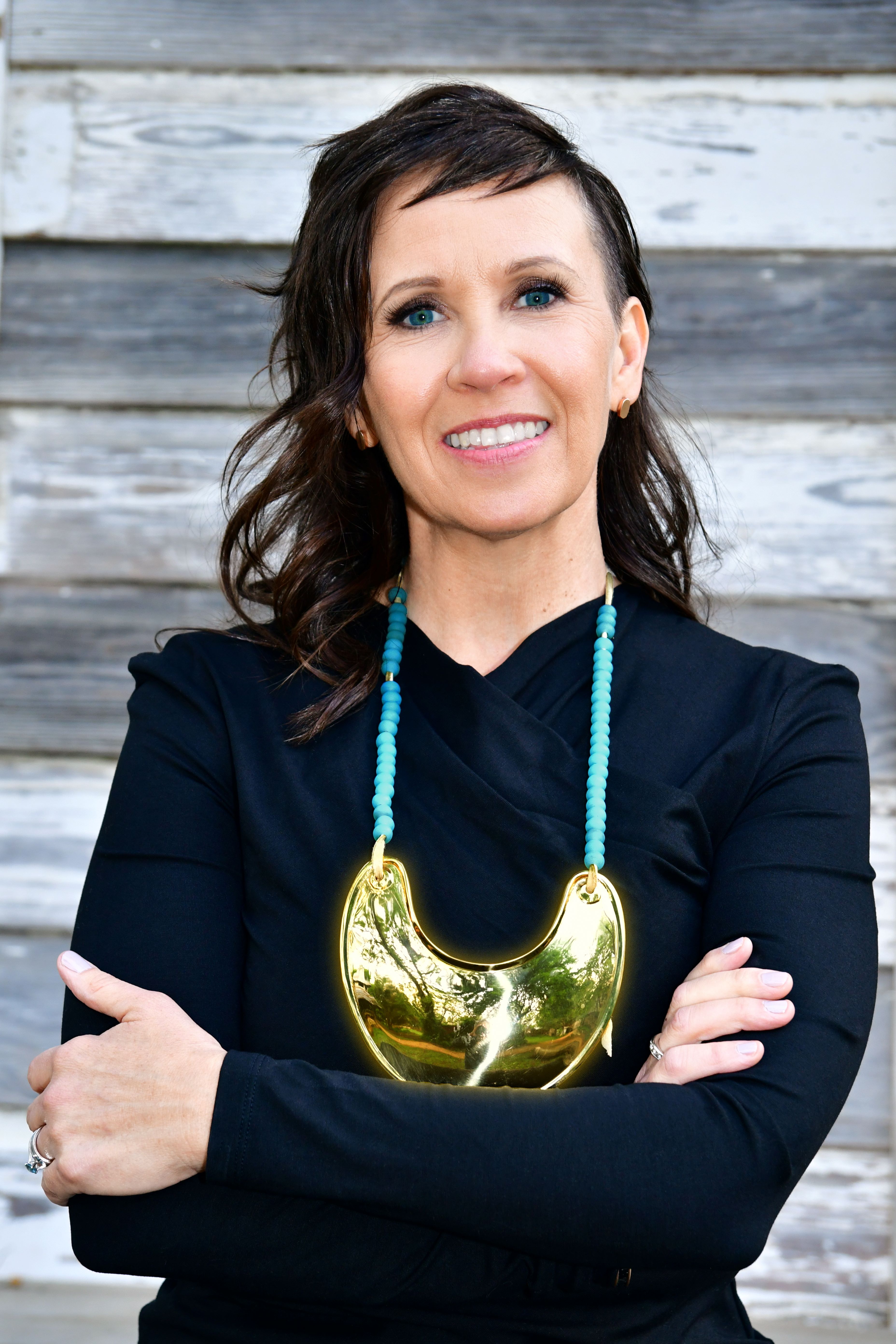 Person wearing a black top and a large gold necklace, standing in front of a wooden background.
