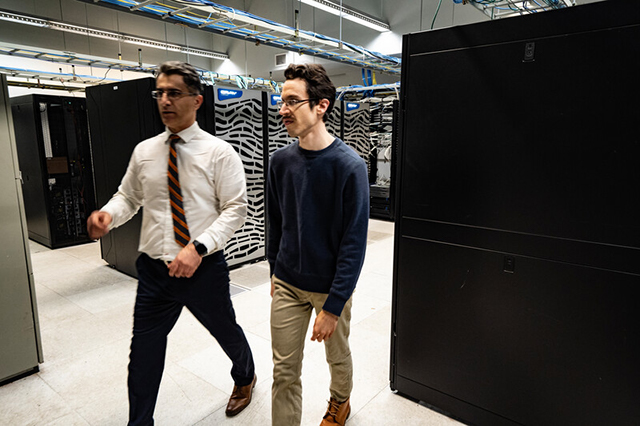 Two men walking through a data center lined with servers. One wears a white shirt and tie, the other a dark sweater. They appear focused and engaged.