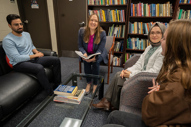 A group of four people sits in a library on a couch and chairs, engaged in a discussion. Bookshelves are filled in the background, creating a scholarly atmosphere.