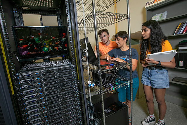 Three people work in a server room, focusing on a monitor displaying colorful graphics. The atmosphere is collaborative and technical.
