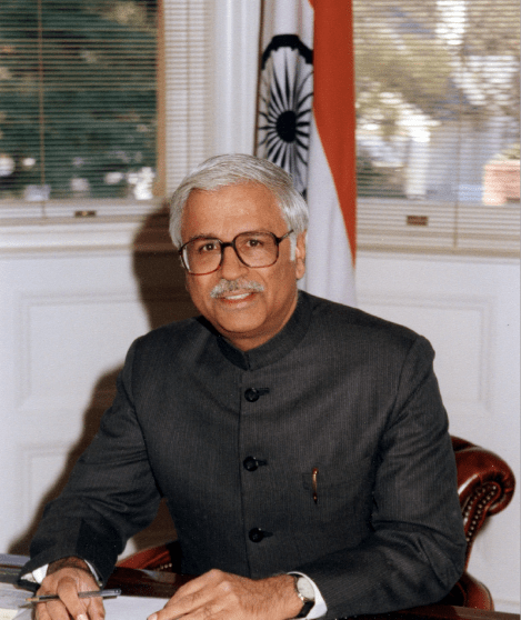 Harsh Basin A professional portrait of a man with grey hair, a mustache, and glasses, wearing a grey Nehru-style jacket. He is seated at a wooden desk, holding a pen over a document. In the background, a white wall with wainscoting is visible next to a window with blinds, and the flag of India stands behind him.