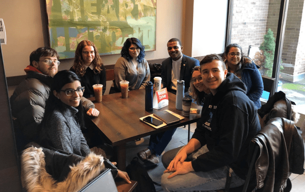A group of eight people are gathered around a dark wooden square table in a brightly lit cafe or common area. They are smiling and looking toward the camera. Seated on the left side: A young man in a tan puffer jacket and a young woman with long dark hair and glasses wearing a grey sweater. Seated along the back: A woman with curly auburn hair in a black top, a woman with dark hair in a grey sweater, and a man in a blue blazer and white collared shirt. Seated on the right side: A young woman in a blue puffer jacket, a woman partially obscured, and a young man in a black hooded sweatshirt and jeans in the foreground. The table holds several iced coffee drinks, a white Starbucks cup, two reusable water bottles, a smartphone, and a small white sign with red text that reads "Blood Drive." A large abstract painting with green and yellow tones hangs on the wall behind them, and a large window to the right shows a brick building and a small evergreen tree outside.