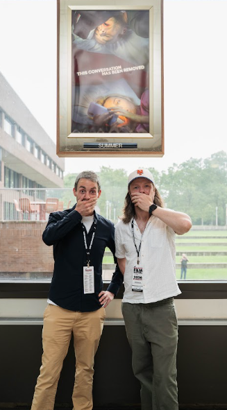 Two men standing in front of poster with hands over mouths.